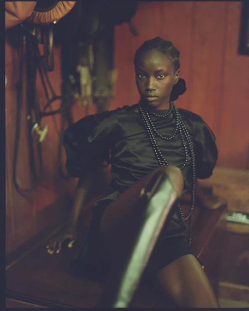 Elegant dark-skinned woman in black dress and leather boots sits on dark surface with horse riding equipment in background.