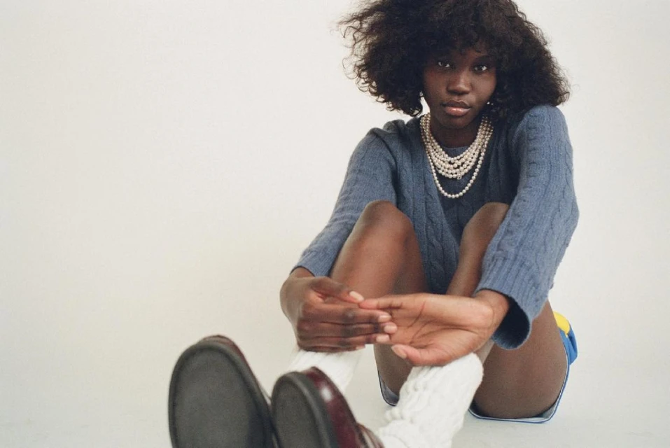Young adult Black woman in a blue sweater and pearl necklaces sitting indoors against a white backdrop, wearing socks and loafers.