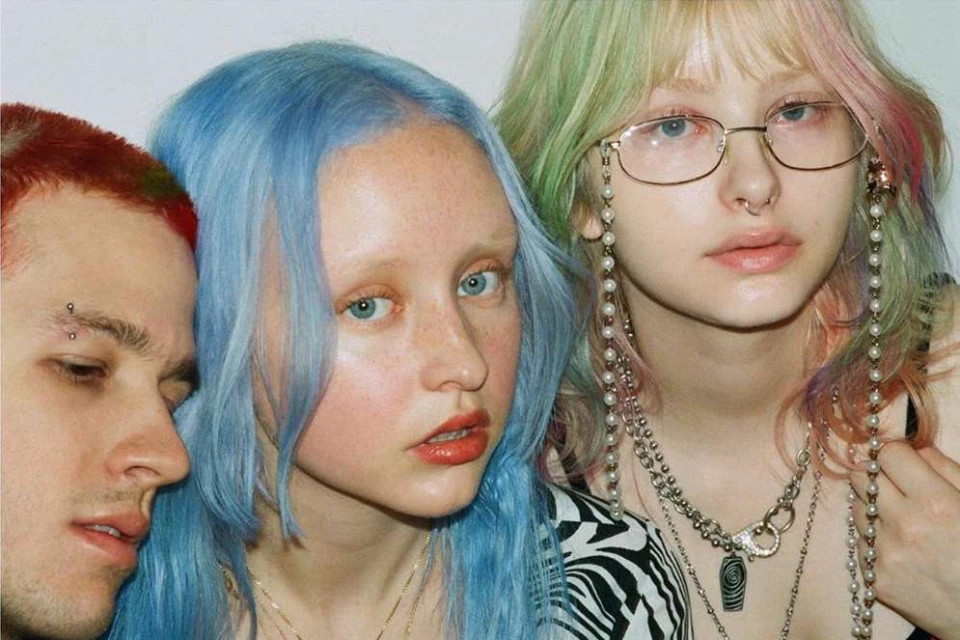 Three young adults pose in a close-up studio shot with colorful hair; red, blue, and green-blonde-pink. A zebra-patterned top is visible.