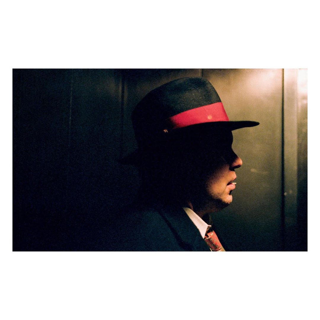 Man in dark suit and fedora with a red band poses in an indoor setting, side profile, mysterious, vintage style, shadowed face.