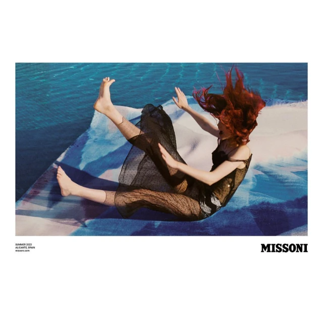 Fashion shot of woman with red hair in a black dress, seated on patterned fabric against a backdrop of blue water, showcasing a Missoni design.