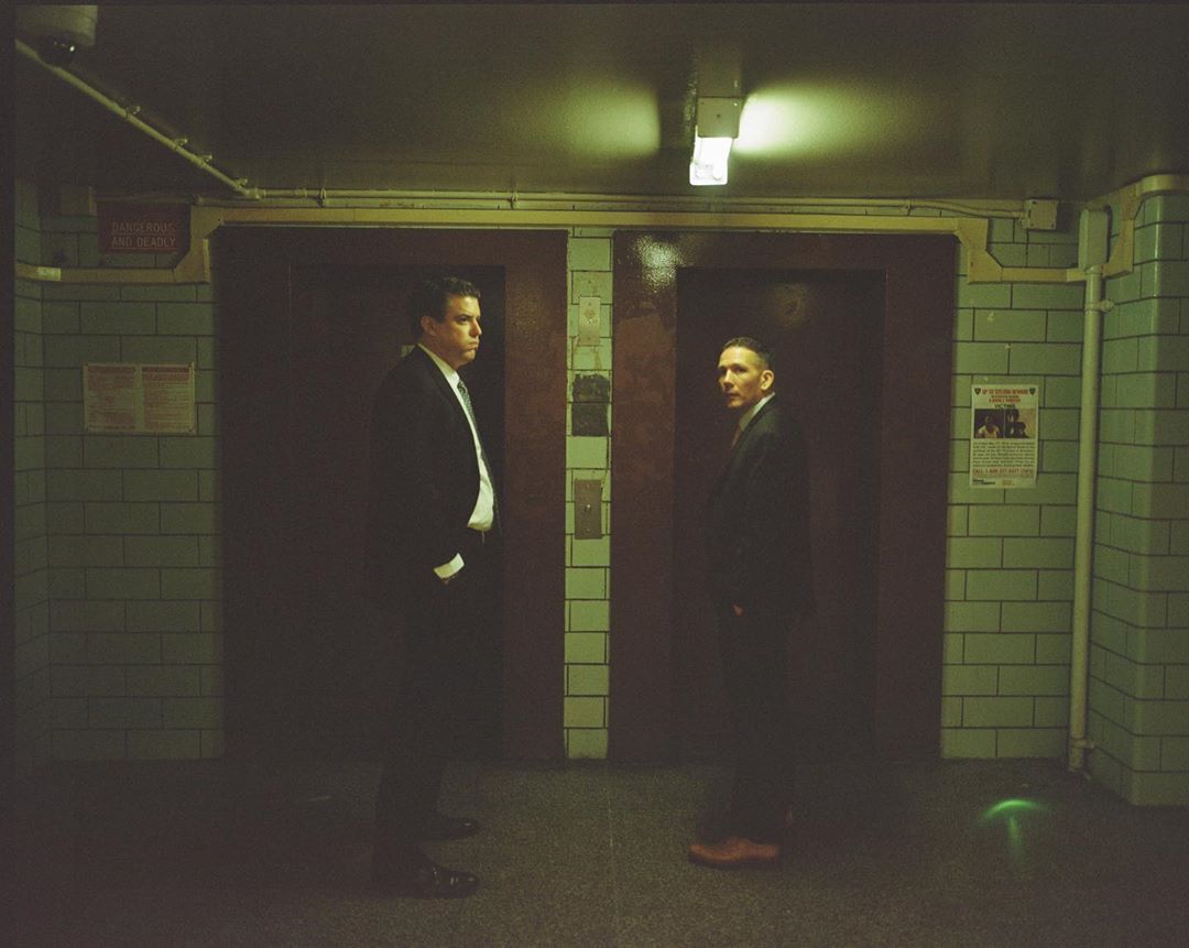 Two men in suits stand in an indoor tiled tunnel under dim lighting, creating a shadowy, formal atmosphere and a sense of depth.