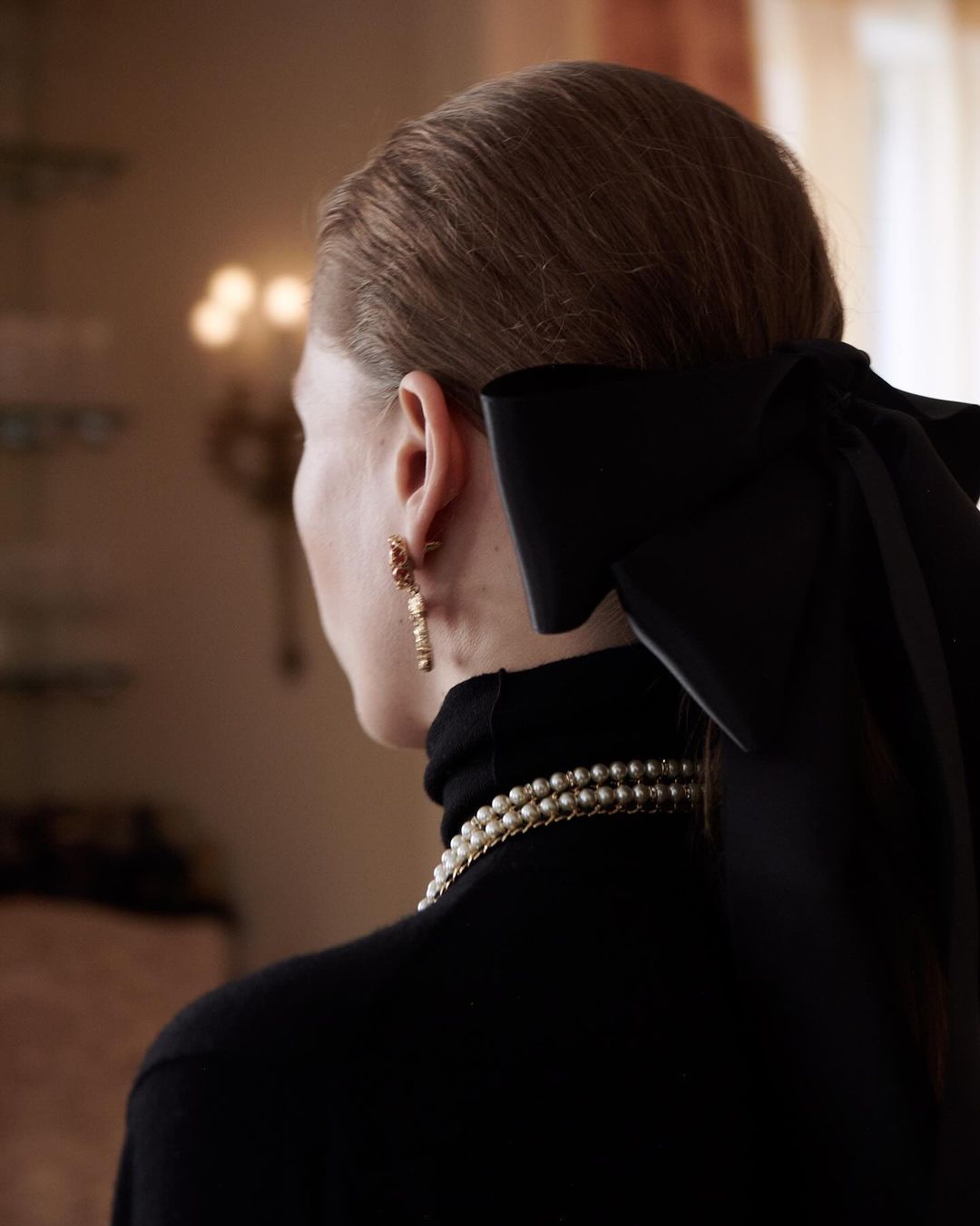 Back view of an adult woman wearing a black turtleneck, pearl necklace and earrings with a large black bow in her hair.