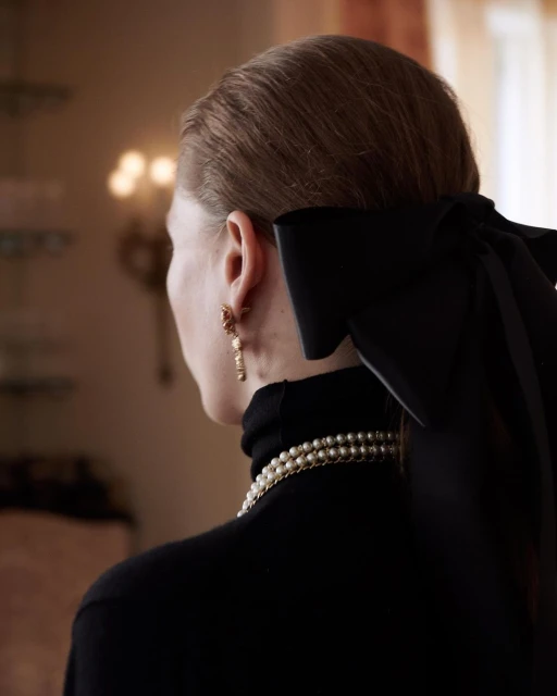 Back view of an adult woman wearing a black turtleneck, pearl necklace and earrings with a large black bow in her hair.