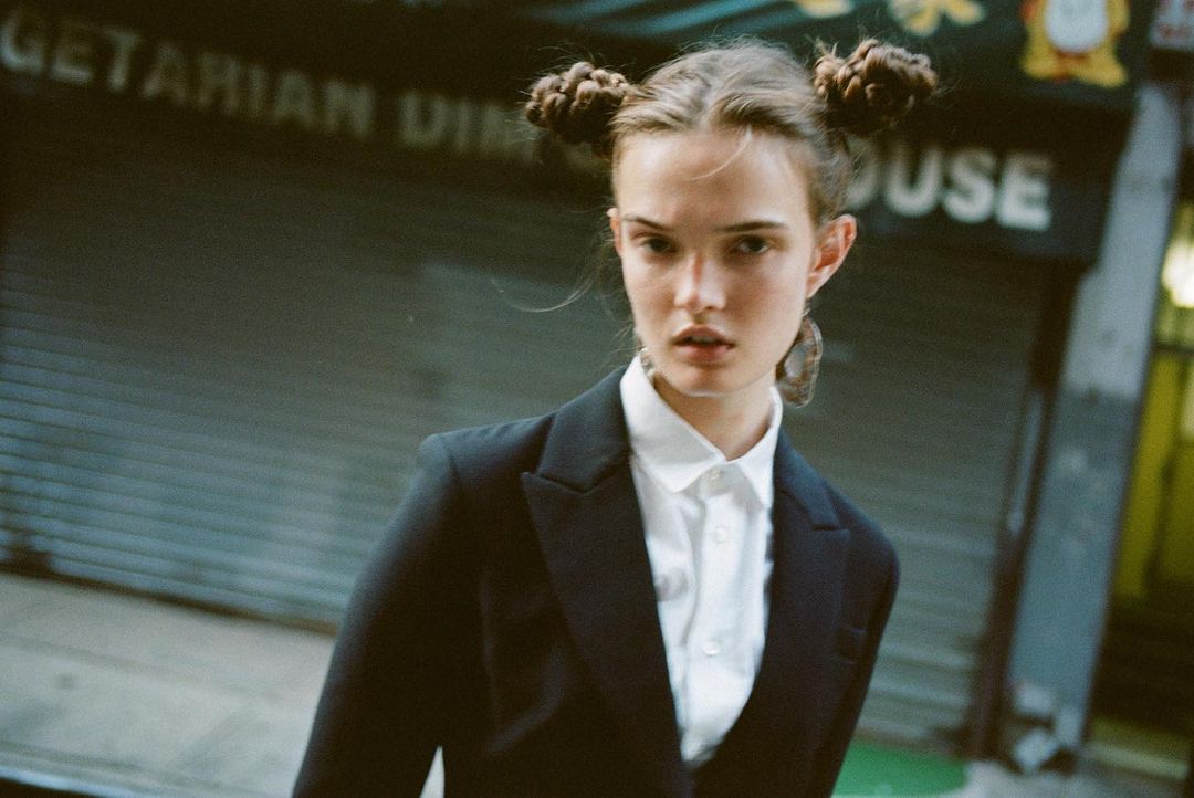 Young adult woman with hair buns in an urban setting. She is wearing a black blazer over a white button-down shirt.