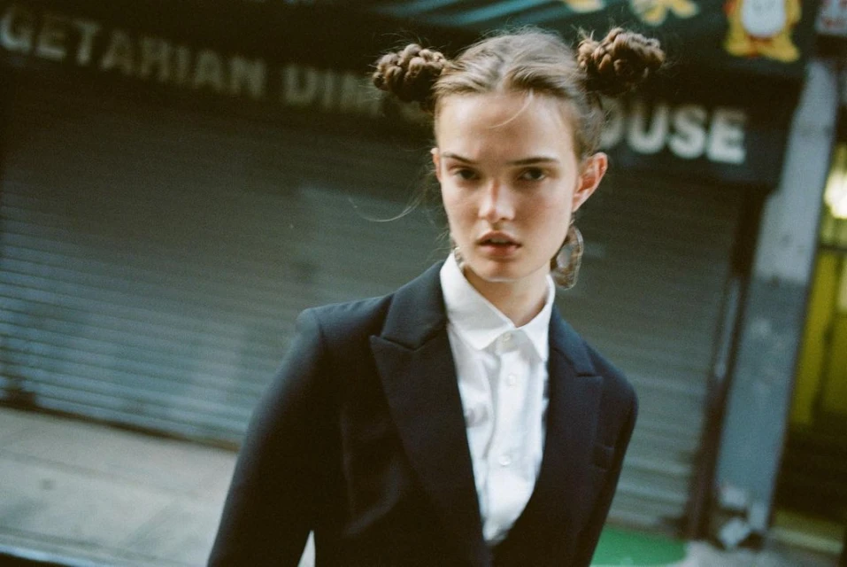 Young adult woman with hair buns in an urban setting. She is wearing a black blazer over a white button-down shirt.