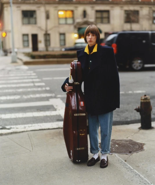 Woman stands on a city street holding a cello case. She wears a black coat, jeans, and loafers, with a city building in the background.