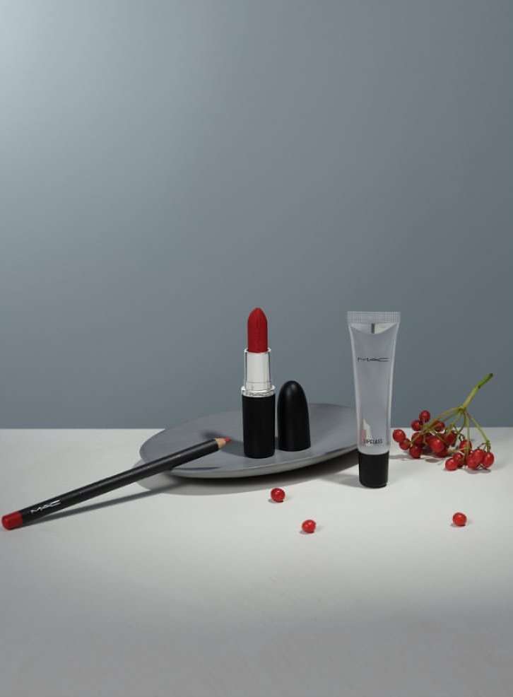 Cosmetic still life: red lipstick, lipliner, and lip gloss arranged on a grey surface with red berries against a grey background.