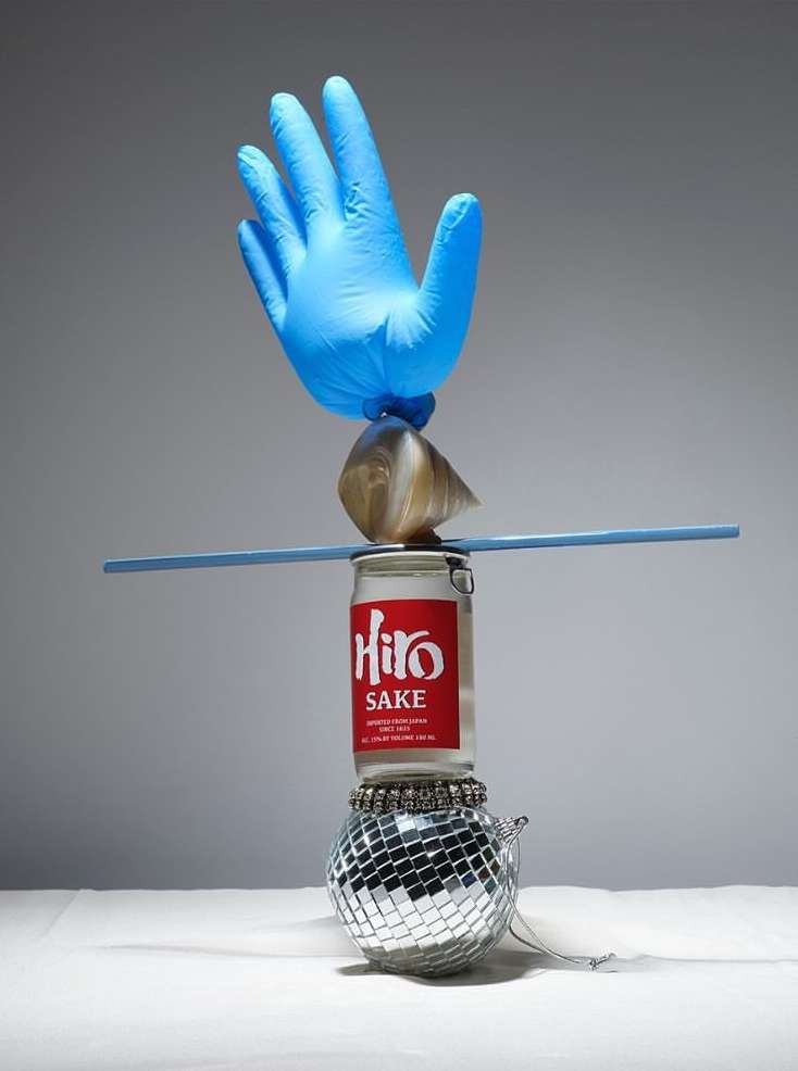 Still life with a blue surgical glove balanced atop a glass jar with sake, a disco ball, and blue chopstick against a grey backdrop.