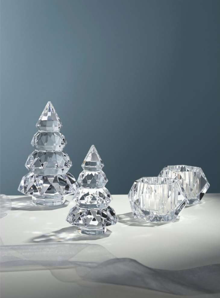 Two crystal Christmas tree figurines with two crystal candle holders on a white table against a blue-grey background, festive and elegant.