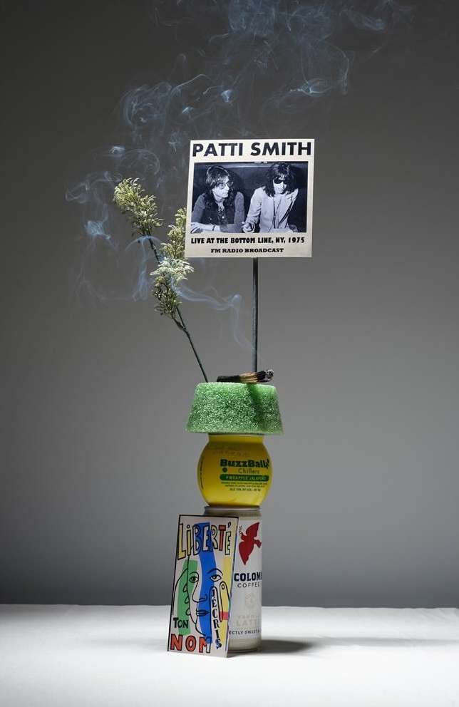 Still life featuring Patti Smith poster, candle, and beverage cans stacked indoors against a gray background, casting a studio lighting.