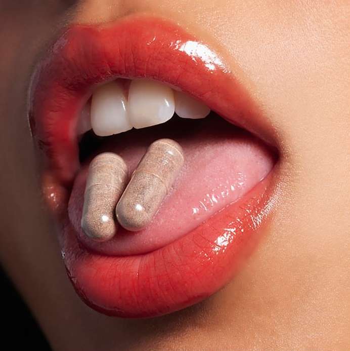Close-up of a woman's mouth open with two capsule pills on her tongue, teeth visible, glossy lipstick, in a high-key lighting setup.