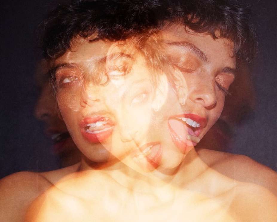 Blurred portrait of a woman with short dark hair and red lipstick, eyes open, and tongue out, against a dark background.