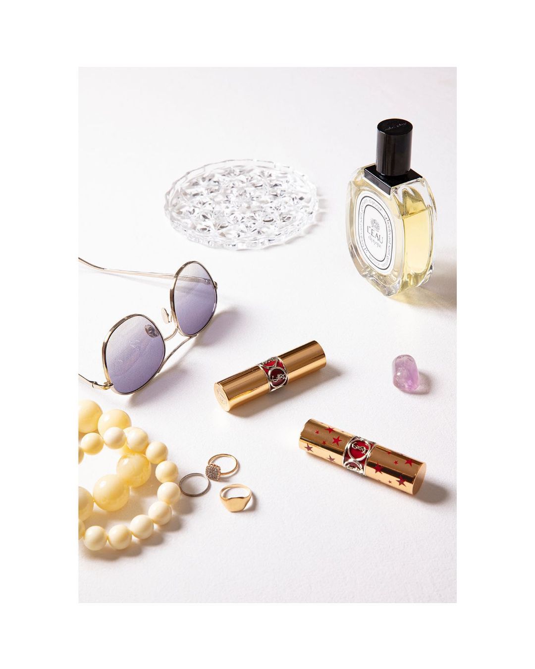 Cosmetic still life: perfume bottle, sunglasses, two lipsticks, rings, beaded necklace, and crystal decoration on a white surface.