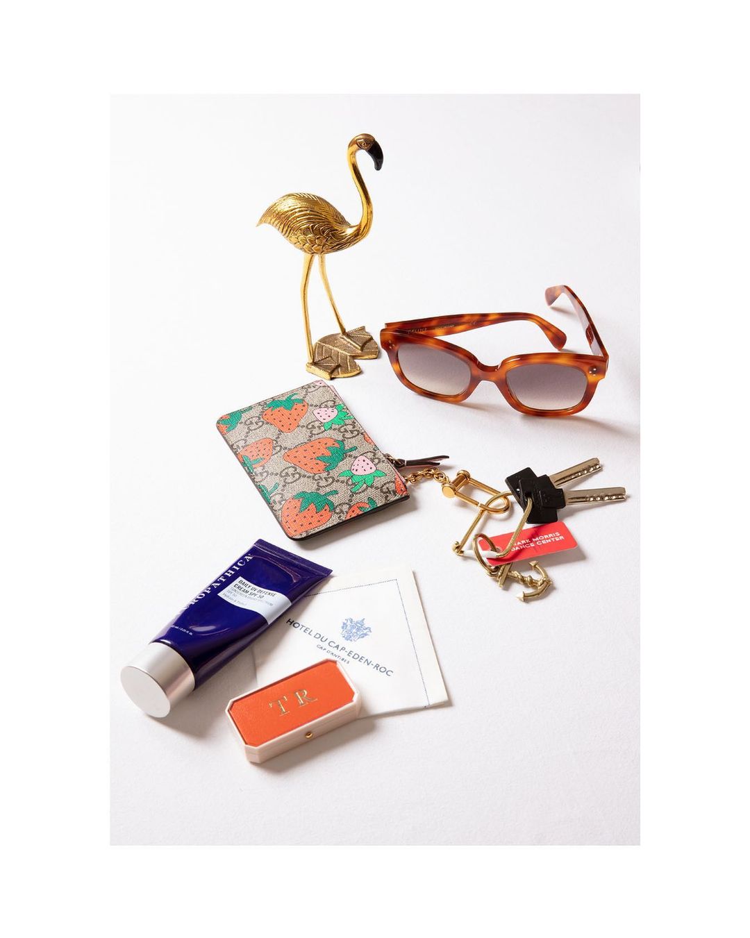 Still life flat lay featuring designer sunglasses, wallet, cosmetics, hotel card, keys, and golden flamingo statue on a white backdrop.