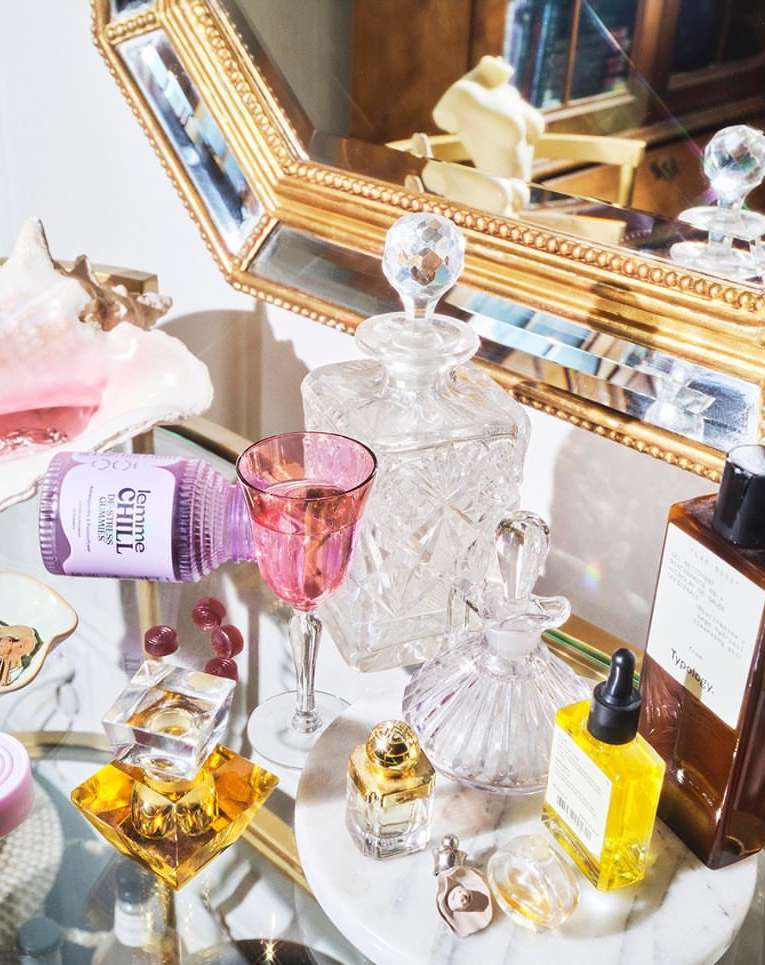 Crystal decanters and glass bottles on a reflective table with marble base, highlighted by pink and yellow hues, and ornate gold framed mirror.