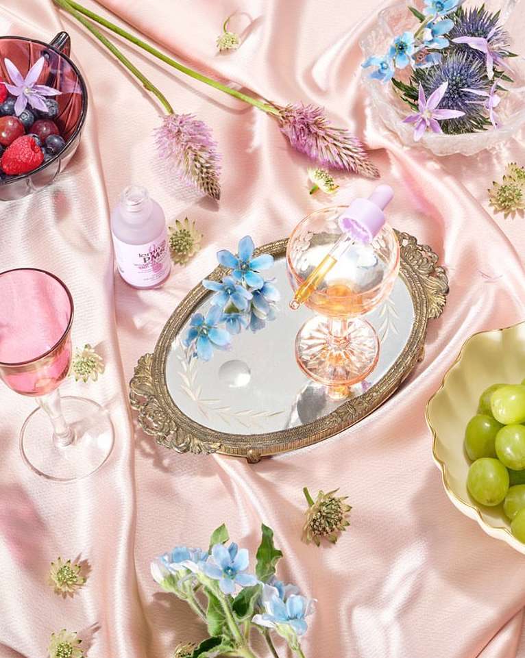 Still life shot of various fruits, glass containers with product, flowers, and a small mirror, arranged on a draped pink satin fabric.