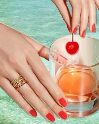 Close-up of hands with red nail polish holding a cherry over a glass of amber drink against sparkling water background
