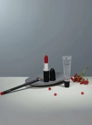 Still life beauty product photography featuring red lipstick, lip pencil, gloss tube, and berries on grey surface.