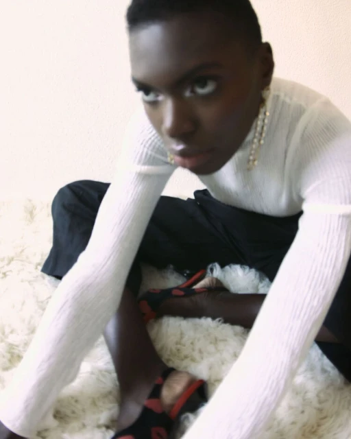 Portrait of a Black woman in a long-sleeved top, black pants, and patterned shoes sitting on a fluffy rug, looking directly at the camera.
