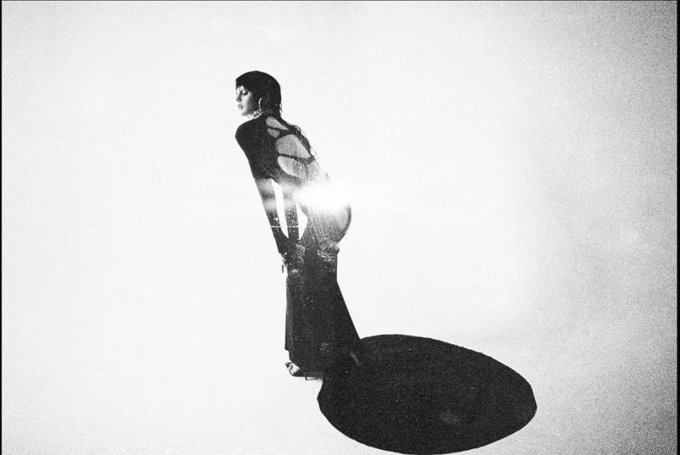 Monochrome portrait of a woman in a black dress bending forward, backlit with a bright light against a bright white backdrop.
