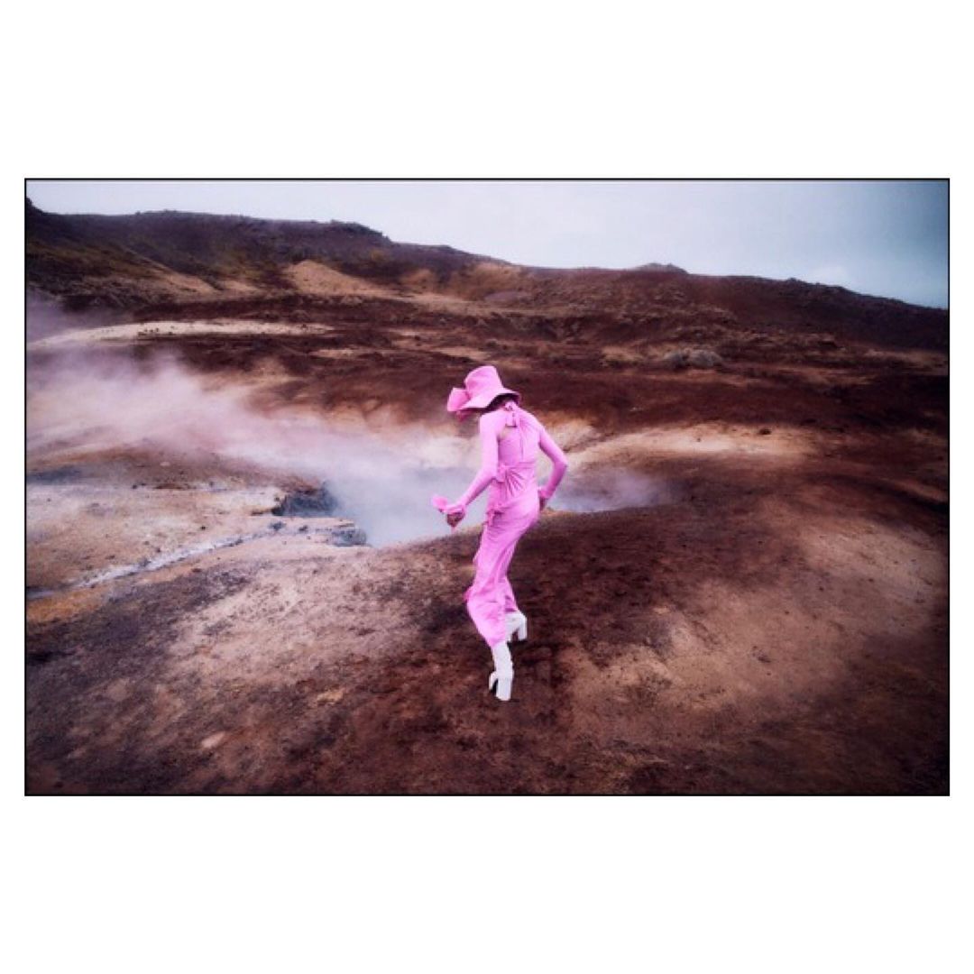 A woman in a pink jumpsuit and hat walks across a brown, volcanic landscape with geothermal vents under a cloudy sky.