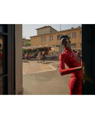 Fashionable woman in red dress and sunglasses peers from doorway onto bustling outdoor market scene in an Italian town.