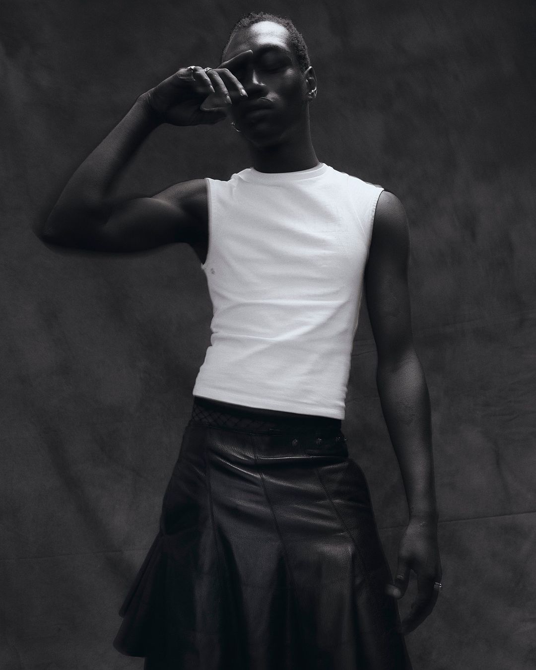 Portrait of a black male in a white sleeveless shirt and black leather skirt, striking a pose against a grey studio background.
