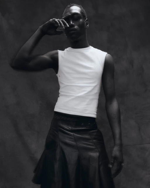 Portrait of a black male in a white sleeveless shirt and black leather skirt, striking a pose against a grey studio background.