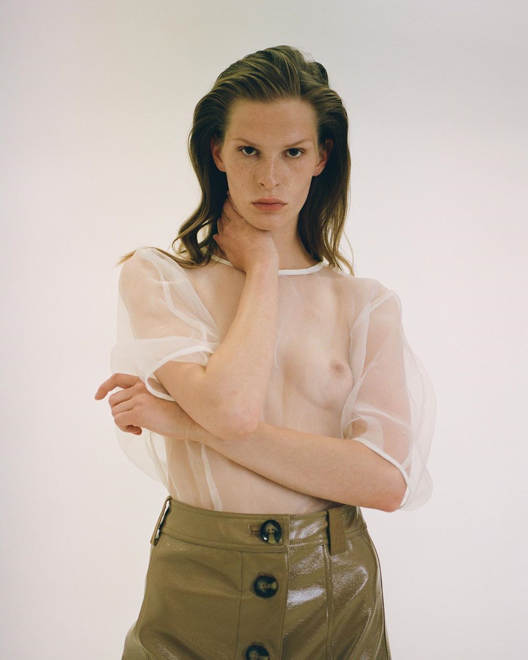 Portrait of a young adult woman with wet hair and freckles, posing in a sheer top and shiny skirt against a clean, white background.