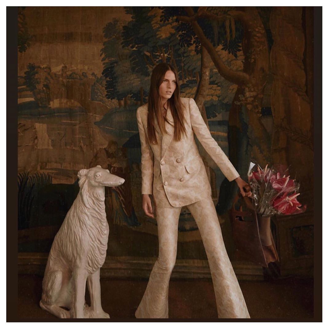 Elegant adult woman poses in a patterned suit with flowers and a dog statue against a vintage scenic tapestry backdrop.