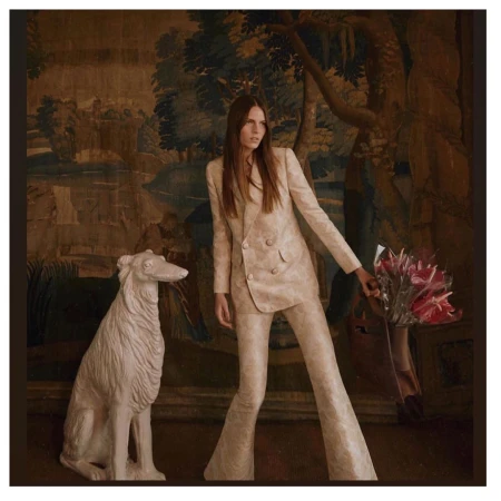 Elegant adult woman poses in a patterned suit with flowers and a dog statue against a vintage scenic tapestry backdrop.