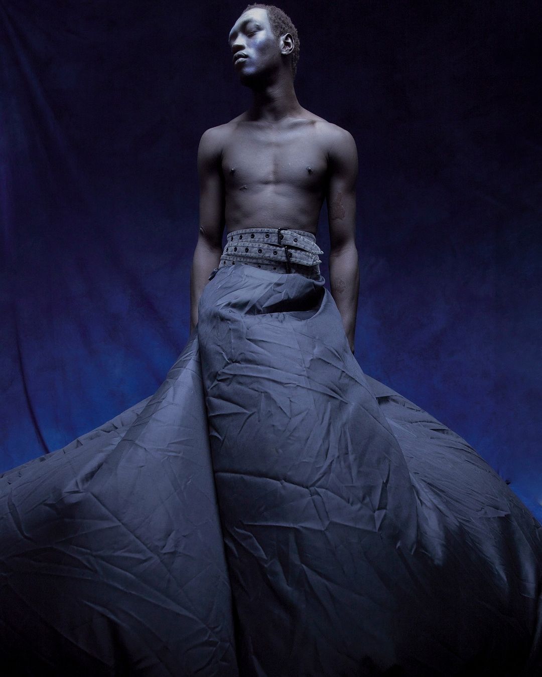 Artistic fashion shot of a topless African American man in a dark blue skirt, standing against a deep blue backdrop in a photo studio.