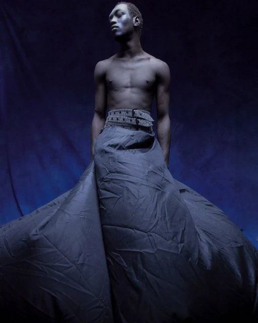 Artistic fashion shot of a topless African American man in a dark blue skirt, standing against a deep blue backdrop in a photo studio.