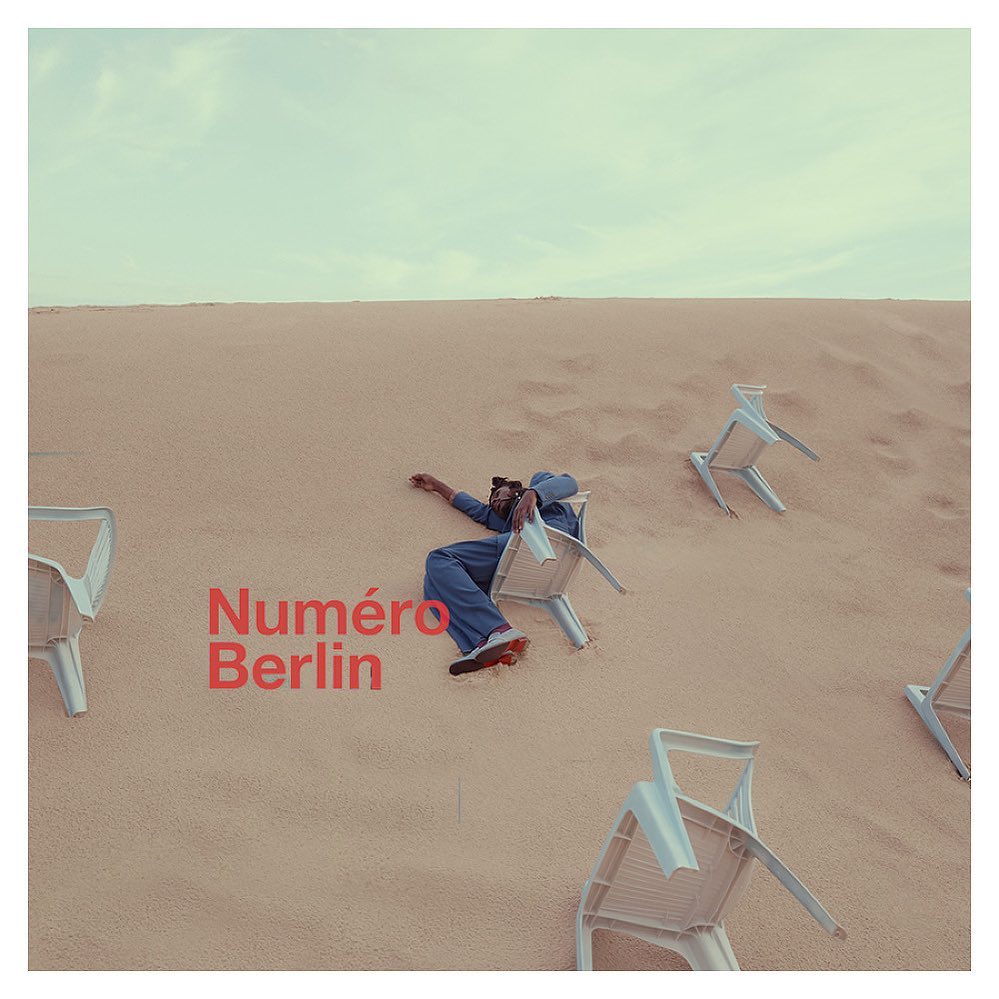 Person in blue jumpsuit rests on a chair in desert landscape with white plastic chairs and sandy dunes beneath a light blue sky.