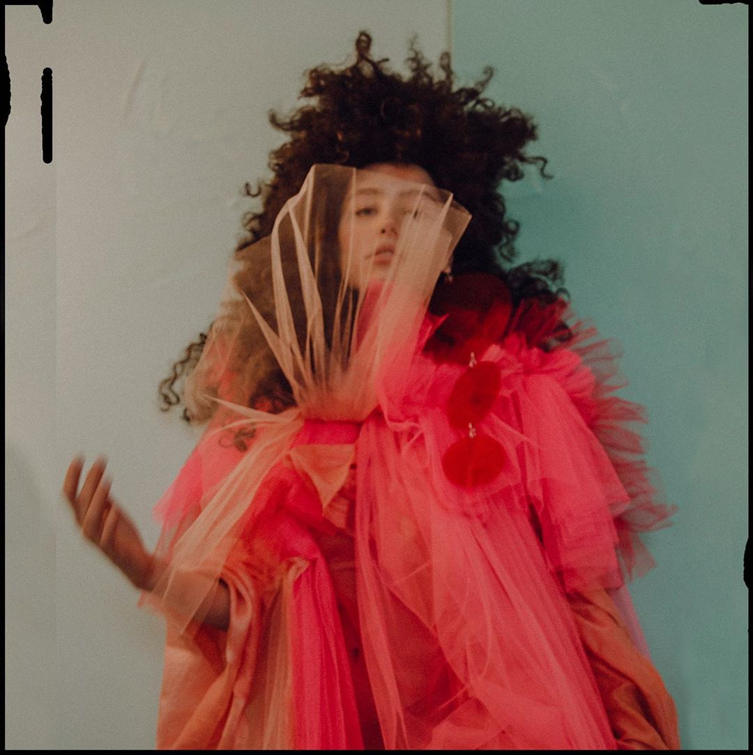 Woman with curly hair wearing a pink tulle gown and a lace veil poses against a pale blue wall in a fashion editorial photograph.