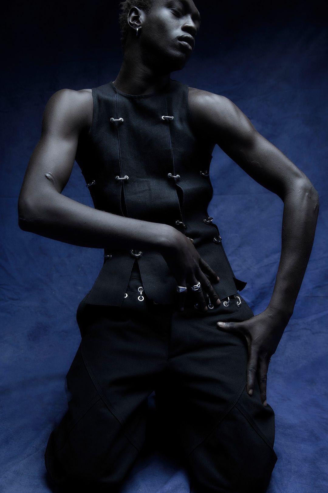 Dark-skinned male fashion model in black vest and pants posing against a dark blue background in a studio setting with artful lighting.
