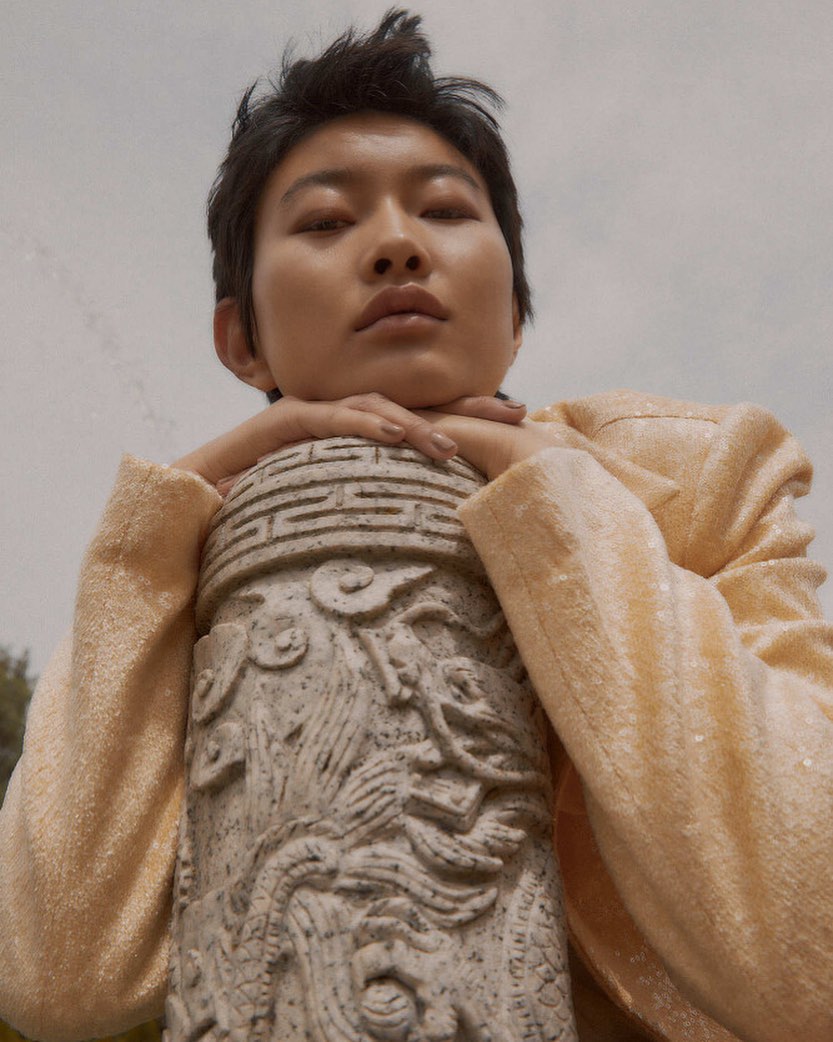 Asian woman with short hair and sequin jacket rests her chin on a carved stone column outdoors under cloudy sky. Fashion editorial shot.