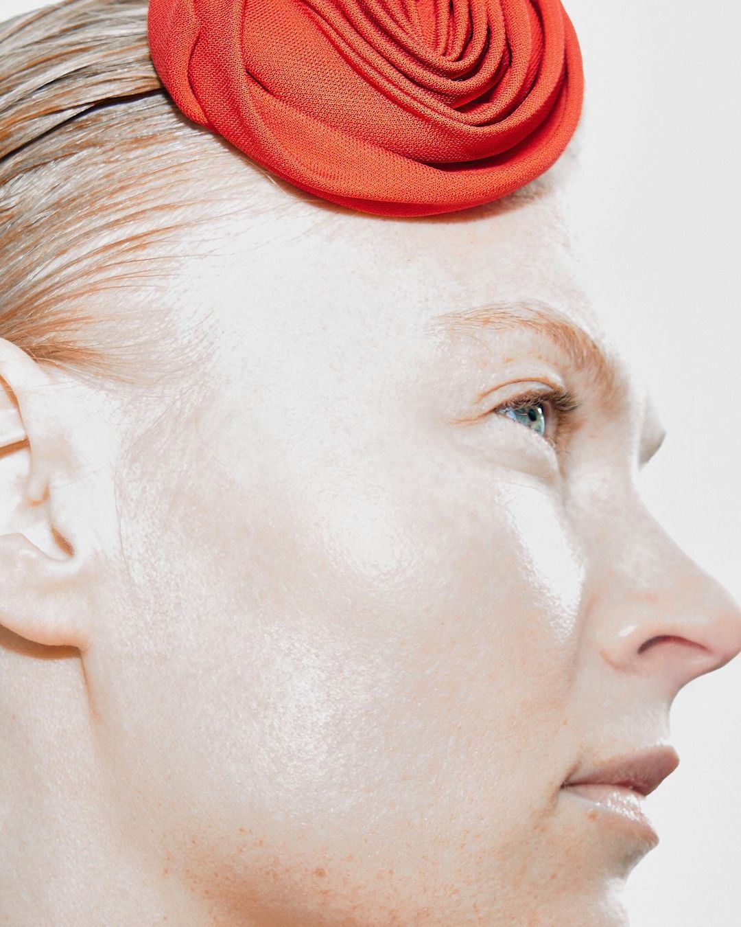 Profile of a woman with fair skin, light eyes and a red flower-like hair accessory against a pale backdrop in soft studio lighting.