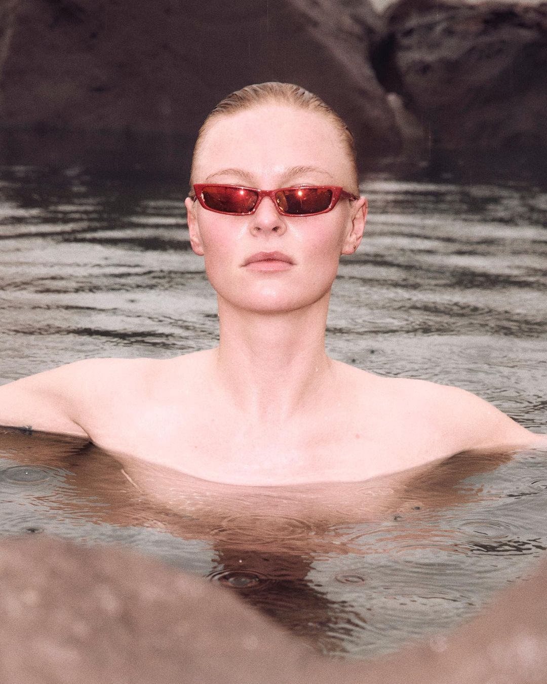 Woman in water wearing sunglasses, looking directly at the camera with neutral expression, wearing red sunglasses, surrounded by grey rocks.