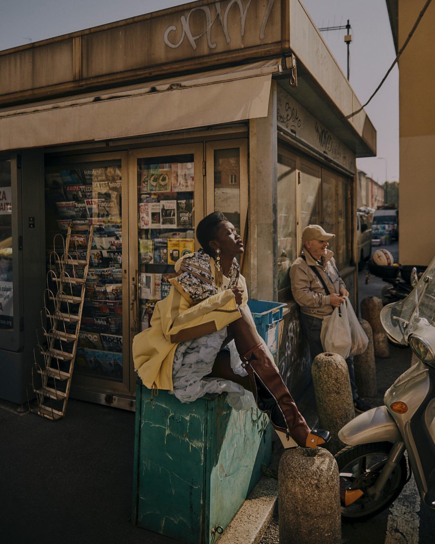 A woman in dress and boots poses on a teal box outdoors next to a newsstand and a scooter, bathed in warm sunlight.