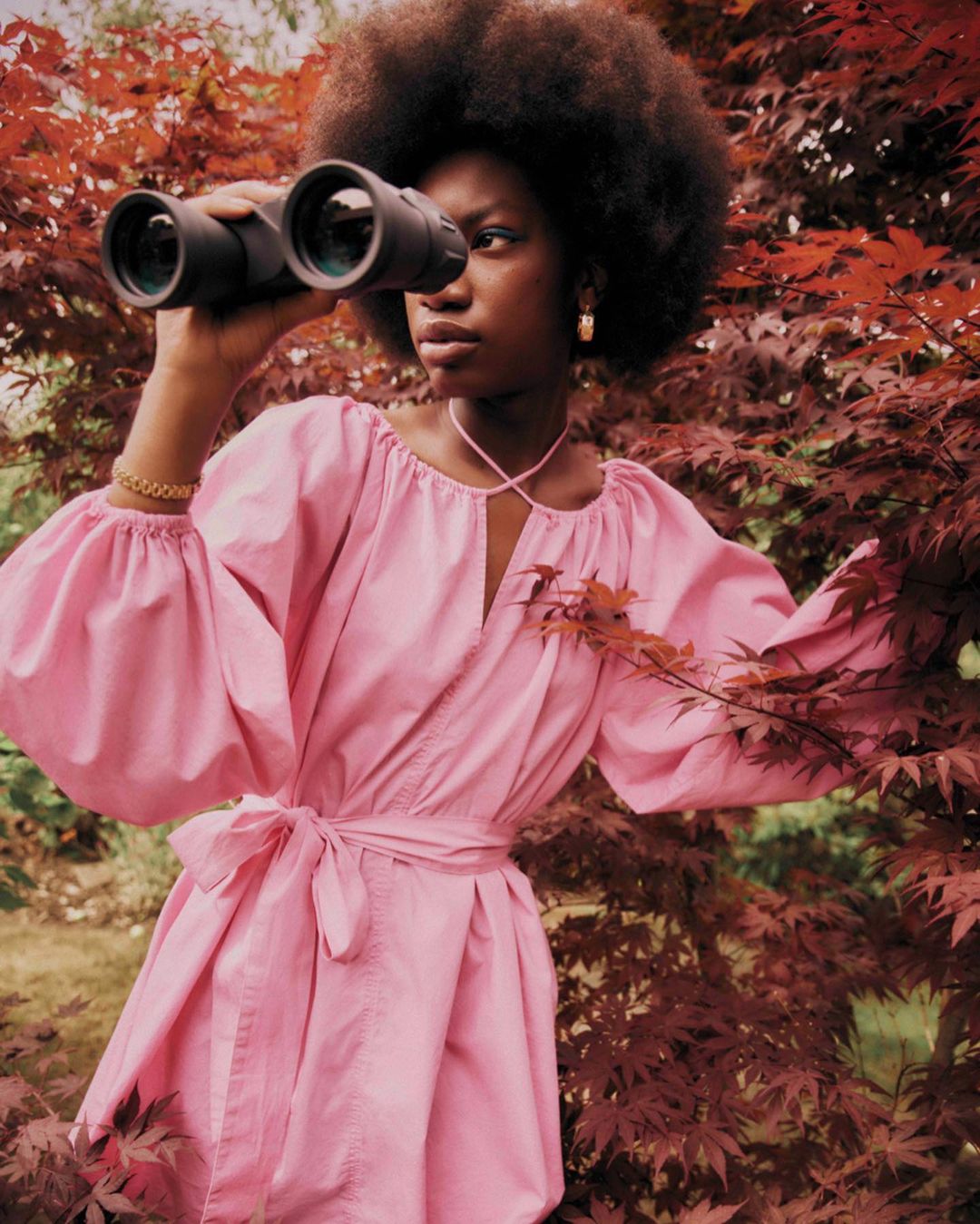 A Black woman stands outside wearing a pink dress and gold hoop earrings, looking through binoculars with red-leafed trees surrounding.