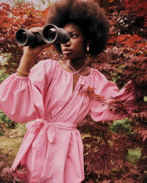 A Black woman stands outside wearing a pink dress and gold hoop earrings, looking through binoculars with red-leafed trees surrounding.