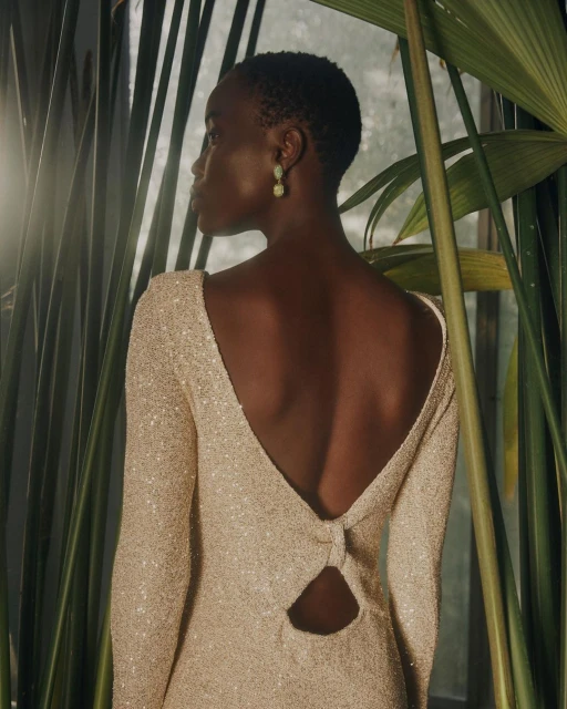 Elegant woman in sparkly dress with open back poses amongst green plants, creating a glamorous and natural aesthetic.
