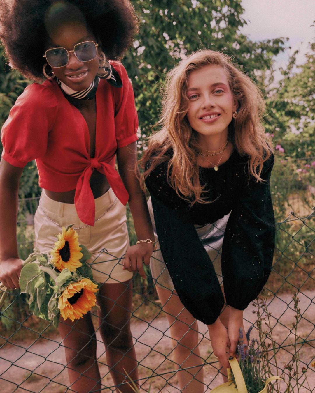 Two young women pose outdoors by a wire fence in sunlight, one in red top holding sunflowers, the other in black top holding watering can.