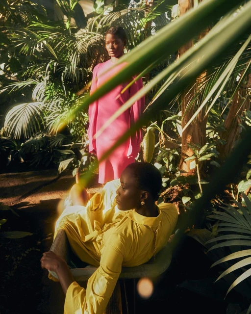 Two women pose in a garden. One woman in a yellow dress sits in a chair in the foreground, another woman stands in a pink dress.