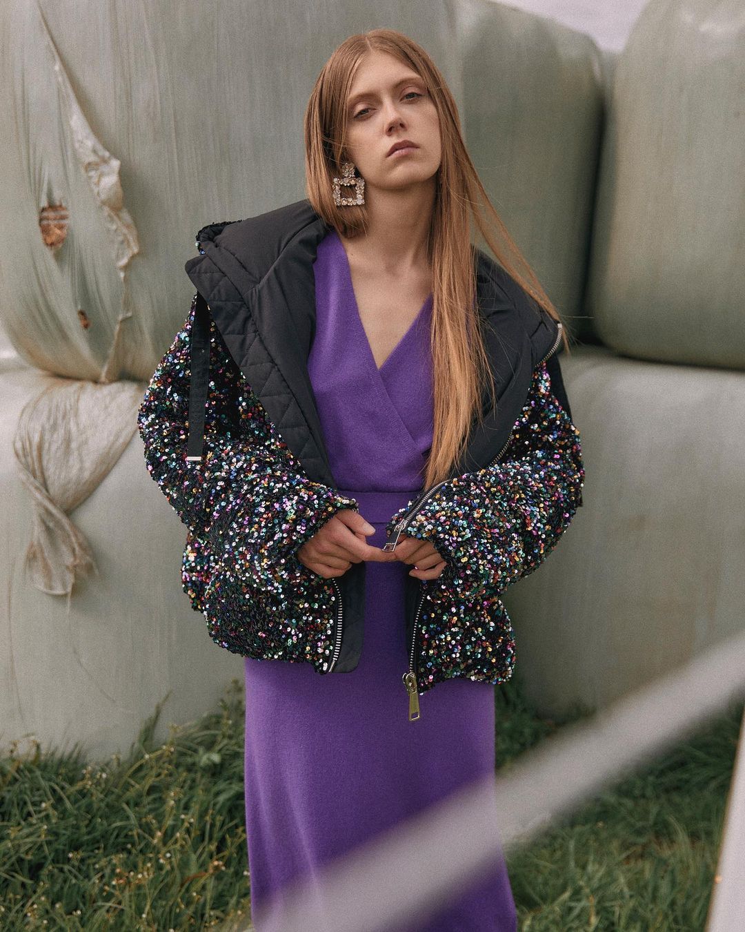 Adult woman with long hair wearing a purple dress and black sequin coat standing outdoors by bales of hay.