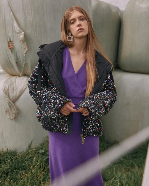 Adult woman with long hair wearing a purple dress and black sequin coat standing outdoors by bales of hay.