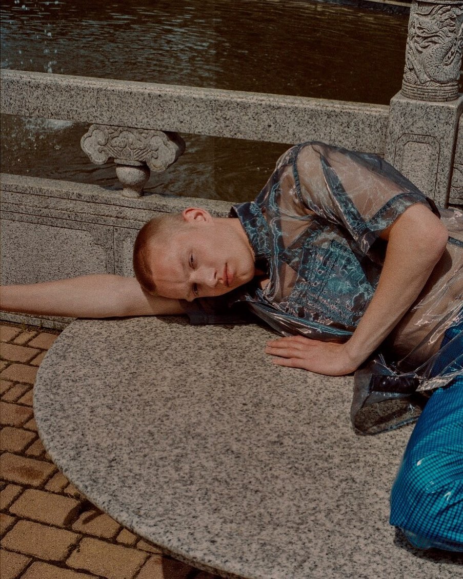 Fair-skinned male with a shaved head lying on a round stone surface outside wearing a sheer blue top and pants with water in the background.