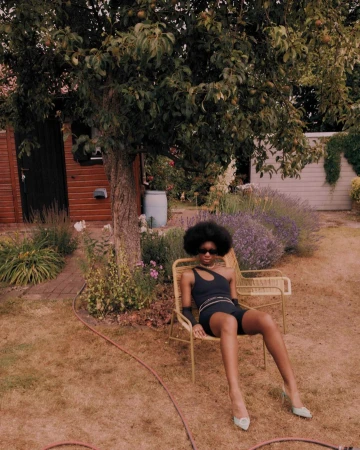 Woman in black shorts and top sitting on a yellow chair in a garden with a large leafy tree and a shed in the background.